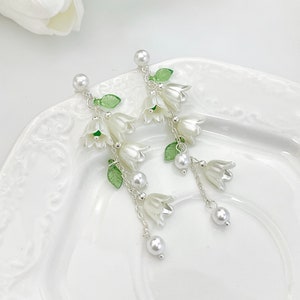 May include: A pair of drop earrings featuring white bell-shaped flower charms, translucent green leaf accents, and small white faux pearl beads suspended from silver-toned metal chains and stud posts with a white pearl top detail.