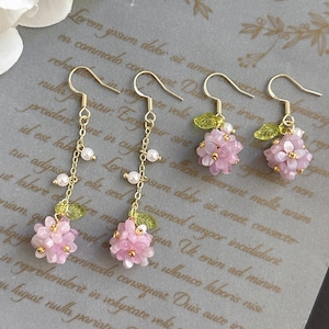 May include: A pair of gold earrings with pink flower charms and pearl accents. The earrings are hanging from a white background with text that reads "Love".