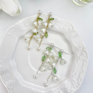May include: A pair of drop earrings featuring white bell-shaped flower charms, small green leaves, and white pearl accents. One earring is suspended on a gold-toned chain, while the other is suspended on a silver-toned chain, both with post backs.