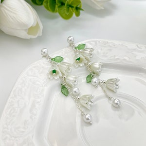 May include: Dangle earrings featuring white bell-shaped flower charms, small green translucent leaves, and round white faux pearl accents suspended from silver-toned chains. The earrings have a post-back closure with a single white pearl stud.