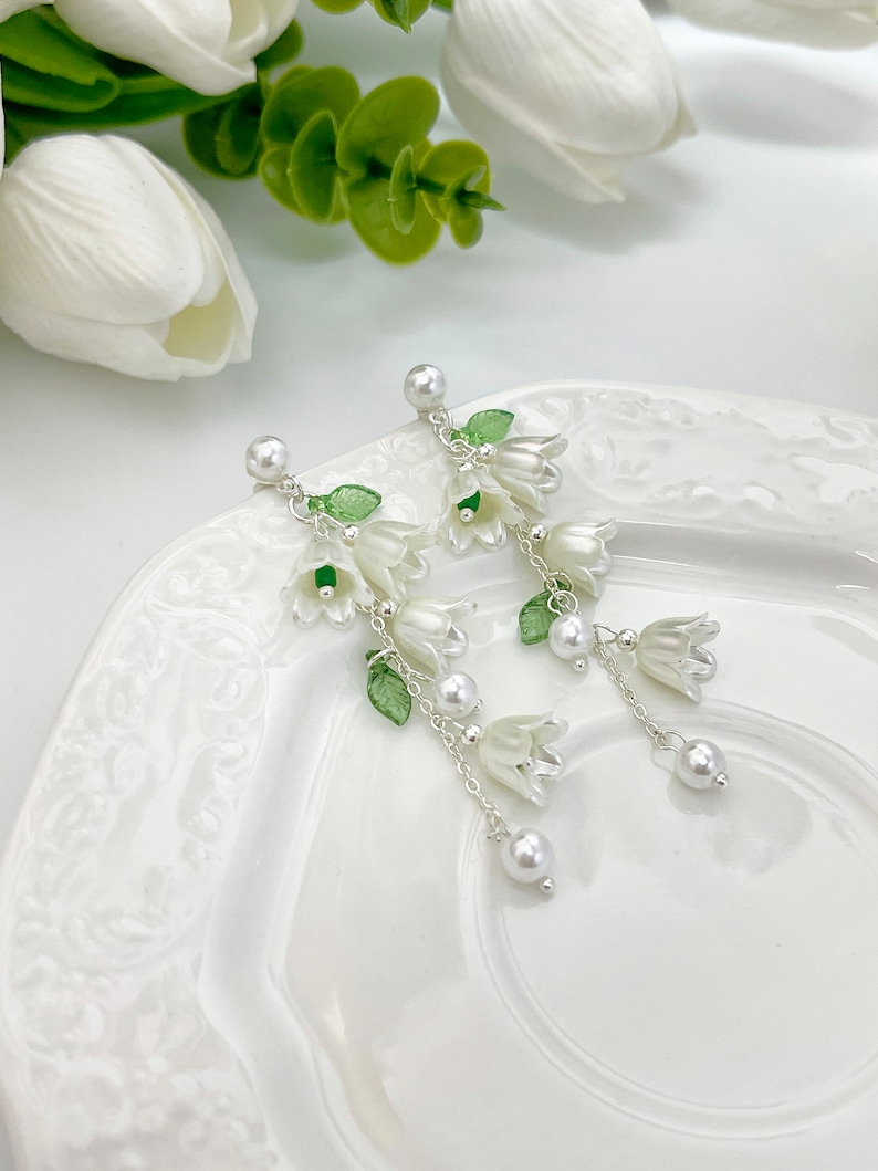 May include: Dangle earrings featuring white bell-shaped flower charms, small green translucent leaves, and round white faux pearl accents suspended from silver-toned chains. The earrings have a post-back closure with a single white pearl stud.