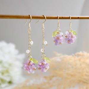 May include: A pair of dangle earrings featuring a cluster of purple flowers and green leaves. The earrings have a gold chain with small white pearls.