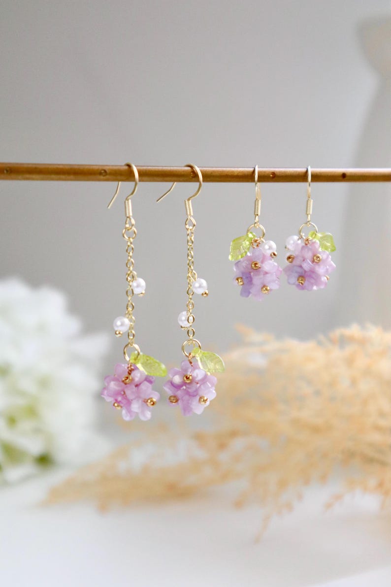 May include: A pair of dangle earrings featuring a cluster of purple flowers and green leaves. The earrings have a gold chain with small white pearls.