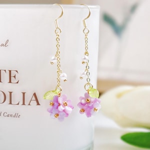 May include: A pair of delicate gold earrings with a cluster of purple and white flowers dangling from each. The flowers are made of small, iridescent beads and are accented with green leaves.