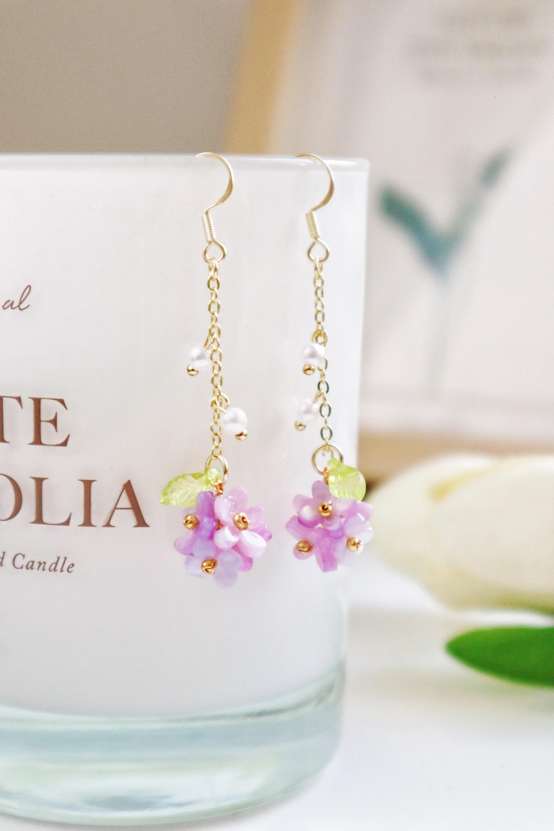 May include: A pair of delicate gold earrings with a cluster of purple and white flowers dangling from each. The flowers are made of small, iridescent beads and are accented with green leaves.