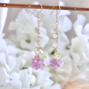 May include: A pair of dangle earrings featuring a cluster of pale purple flowers and green leaves at the bottom. The earrings are crafted with gold chain and white pearl accents.