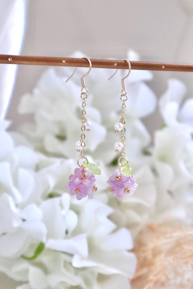 May include: A pair of dangle earrings featuring a cluster of pale purple flowers and green leaves at the bottom. The earrings are crafted with gold chain and white pearl accents.