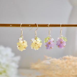 May include: Three pairs of floral earrings with gold hooks. The earrings feature clusters of small, pastel-coloured flowers with green leaves. The flowers are yellow, purple, and light purple.