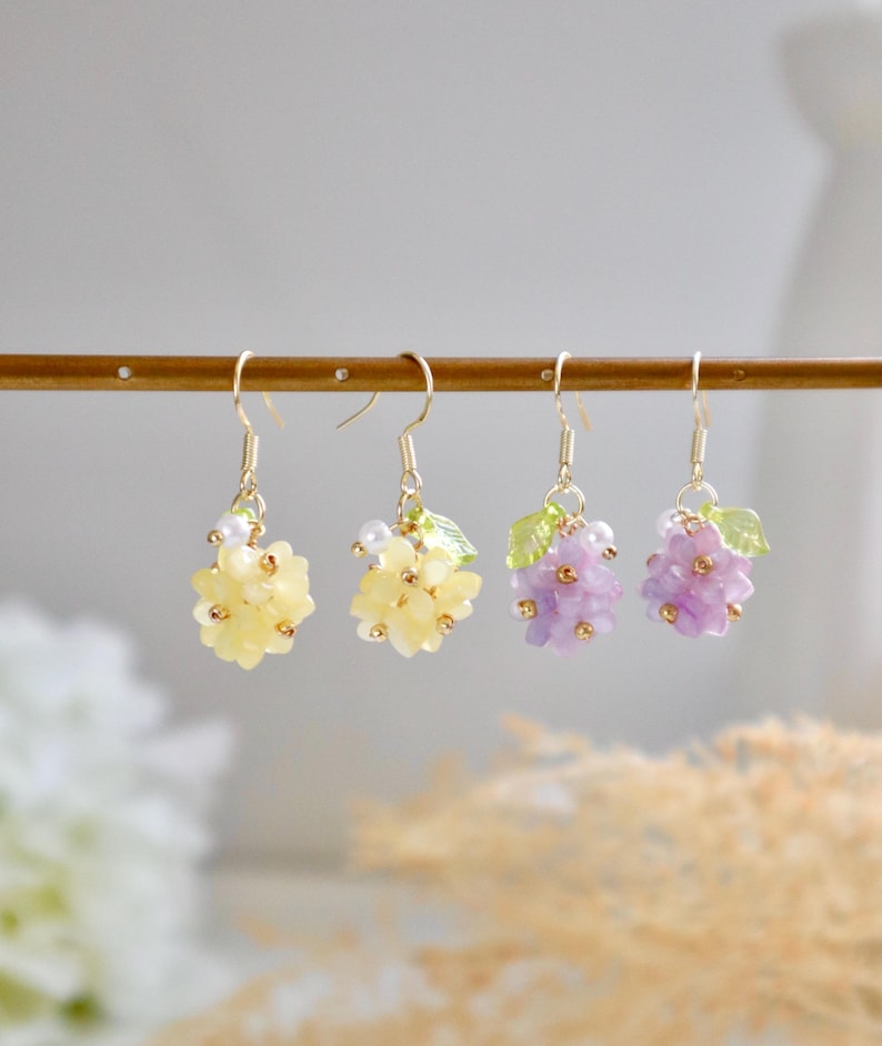 May include: Three pairs of floral earrings with gold hooks. The earrings feature clusters of small, pastel-coloured flowers with green leaves. The flowers are yellow, purple, and light purple.