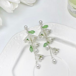 May include: A pair of drop earrings featuring white bell-shaped flower charms, small green leaves, and silver-toned chains. Each earring is accented with white faux pearl beads at the top and bottom, suspended from a silver-toned post backing.