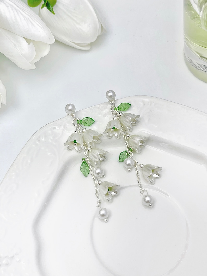 May include: A pair of drop earrings featuring white bell-shaped flower charms, small green leaves, and silver-toned chains. Each earring is accented with white faux pearl beads at the top and bottom, suspended from a silver-toned post backing.