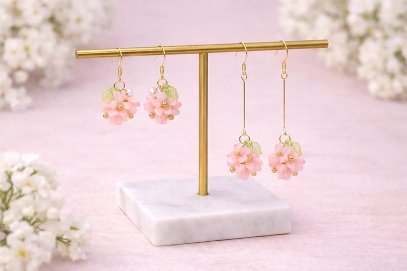 May include: A display of four pairs of pink flower earrings on a gold-colored earring stand. The earrings have a cluster of pink flowers with gold accents and green leaves. The stand is on a white marble base.