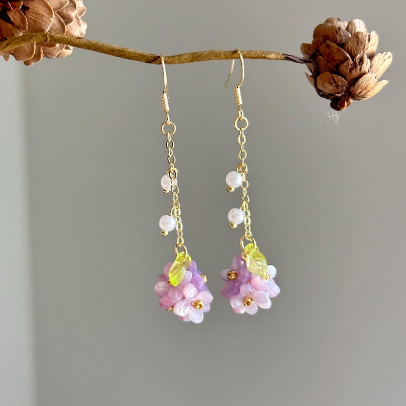 May include: A pair of gold dangle earrings with a cluster of pink and purple flowers and green leaves at the bottom. The earrings have a chain with small white beads.