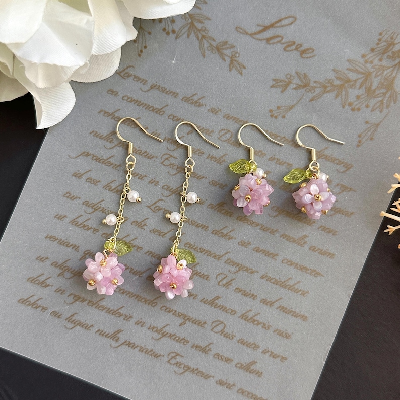 May include: A pair of gold earrings with pink flower charms and pearl accents. The earrings are hanging from a white background with text that reads "Love".