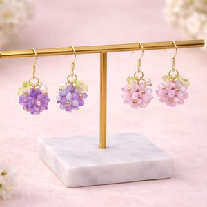 May include: Earrings displayed on a gold stand. Two pairs of earrings are visible, each with a cluster of beads resembling grapes. One pair is purple, the other is pink. The earrings have gold hooks and small green leaf accents.