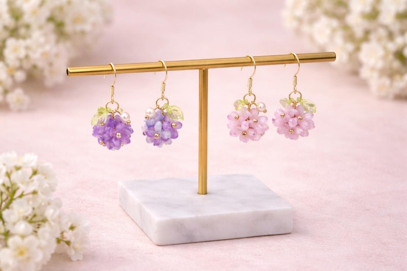 May include: Earrings displayed on a gold stand. Two pairs of earrings are visible, each with a cluster of beads resembling grapes. One pair is purple, the other is pink. The earrings have gold hooks and small green leaf accents.