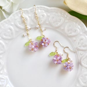May include: A pair of delicate earrings featuring clusters of purple and white flowers with green leaves dangling from gold chains.
