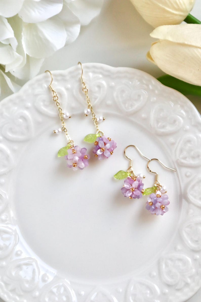 May include: A pair of delicate earrings featuring clusters of purple and white flowers with green leaves dangling from gold chains.