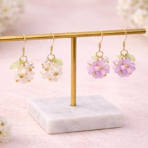 May include: Earrings displayed on a gold stand. Two pairs of beaded earrings are shown, one in white and the other in lavender. Each earring has a cluster of beads and small green leaf accents. The stand is on a marble base.