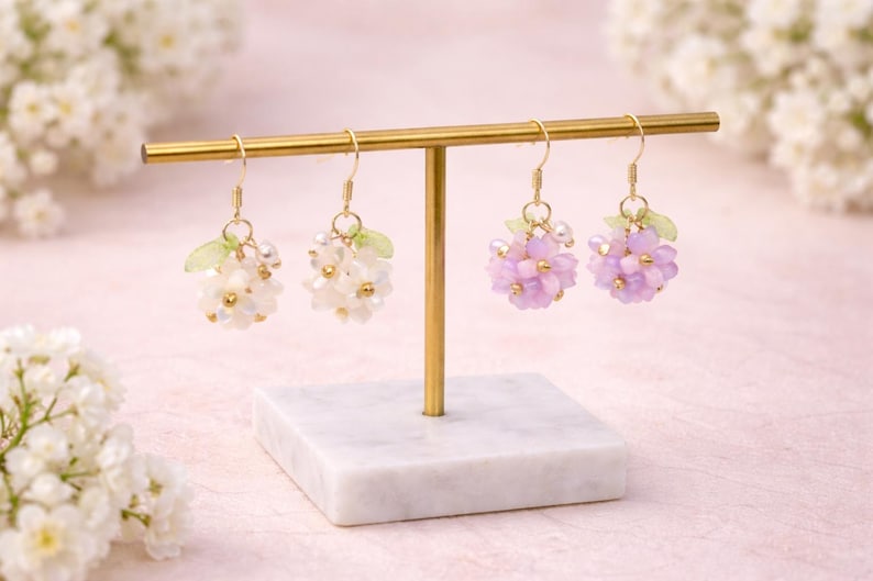 May include: Earrings displayed on a gold stand. Two pairs of beaded earrings are shown, one in white and the other in lavender. Each earring has a cluster of beads and small green leaf accents. The stand is on a marble base.