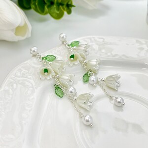 May include: Dangle earrings featuring white bell-shaped flower charms, small green leaves, and white faux pearl beads suspended from silver-toned metal chains. The earrings have a post-back closure with a single white faux pearl at the top.