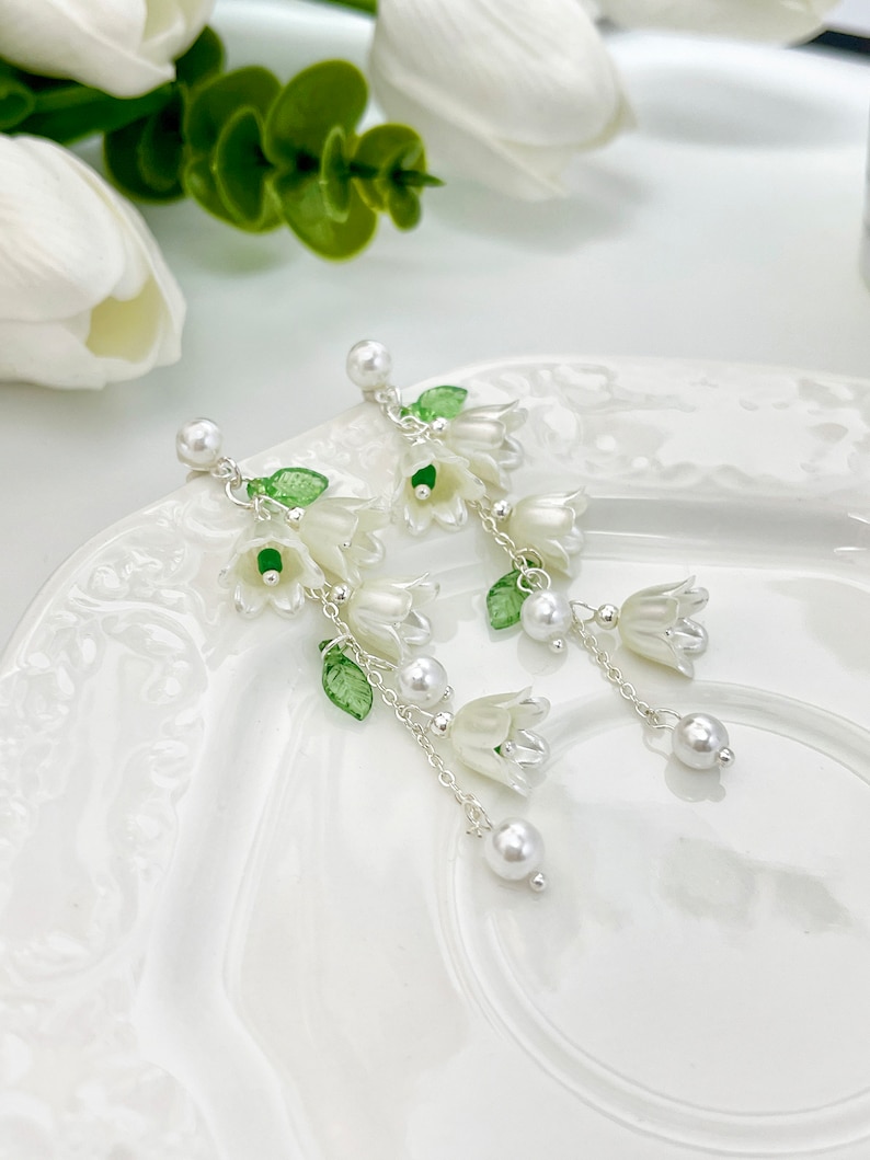 May include: Dangle earrings featuring white bell-shaped flower charms, small green leaves, and white faux pearl beads suspended from silver-toned metal chains. The earrings have a post-back closure with a single white faux pearl at the top.