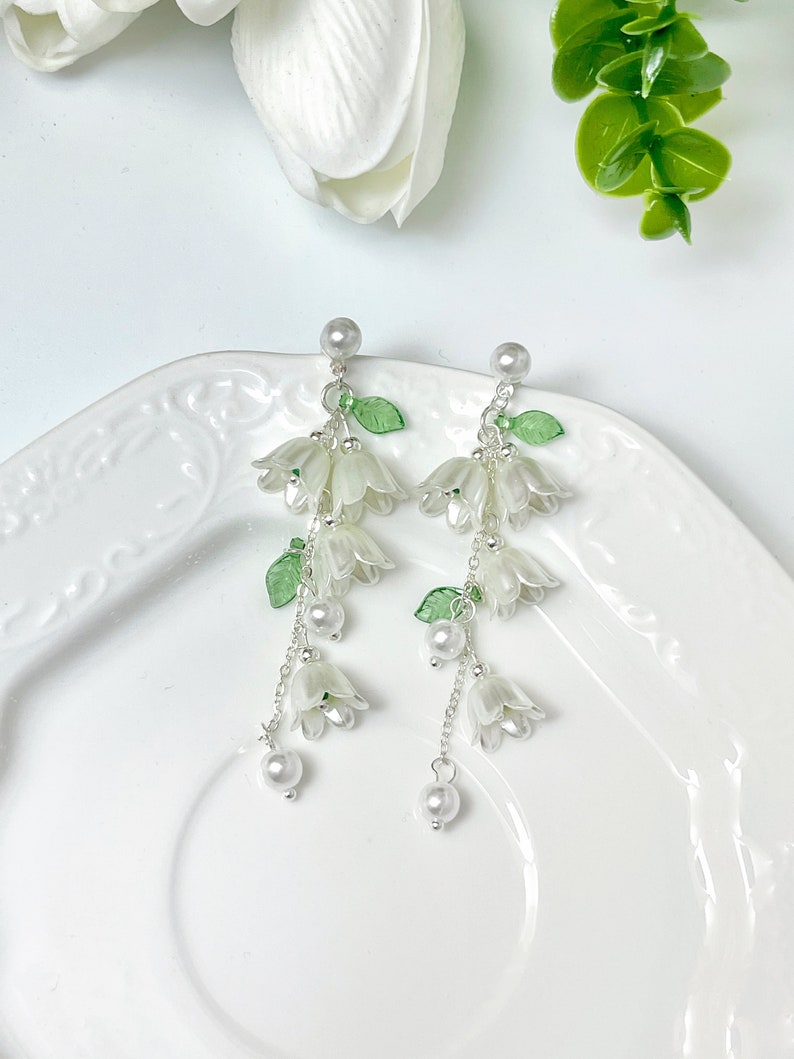 May include: A pair of drop earrings featuring translucent white bell-shaped flower charms, small green leaf accents, and round white faux pearl beads suspended from silver-toned metal chains with a stud post closure at the top.