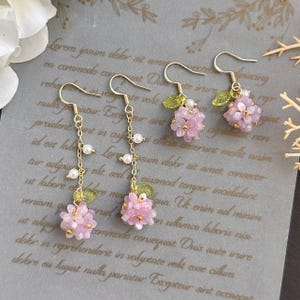 May include: A pair of gold earrings with pink flower charms and pearl accents. The earrings are hanging from a gold chain with a small green leaf charm.