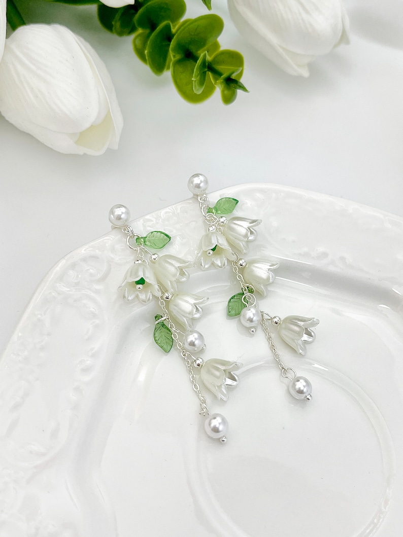May include: A pair of drop earrings featuring white bell-shaped flower charms, translucent green leaf accents, and small white faux pearl beads suspended from silver-toned metal chains and stud posts with a white pearl top detail.