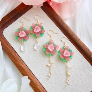 Peut inclure: Trois paires de boucles d'oreilles florales avec des motifs de fleurs de lotus roses et vertes. Les boucles d'oreilles ont des crochets dorés et des accents de cristaux clairs et de perles. Les boucles d'oreilles sont présentées sur un plateau en bois.