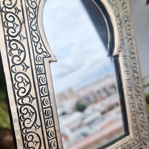 May include: An ornate silver-coloured mirror with a decorative, intricate, carved frame. The mirror has an arched top and reflects a blurred image of a city.