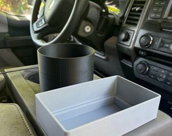 Op de afbeelding: Een grijze plastic opbergbak en een zwarte bekerhouder voor een auto. De bak is rechthoekig en heeft een rand rondom. De bekerhouder is cilindrisch en heeft een getextureerd oppervlak.