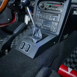 May include: The interior of a car with a black leather gear shift boot and a silver gear shift knob. The car has black carpet and a black dashboard with a radio and climate controls. There are two black window switches on the centre console.