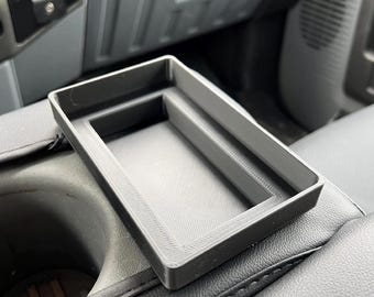Op de afbeelding: Een zwarte rechthoekige console-organizer, waarschijnlijk voor een voertuig, ligt op een zwart leren oppervlak. De organizer heeft een gestructureerd interieur en een verhoogde rand. De achtergrond toont het interieur van een voertuig, inclusief een bekerhouder en dashboard.