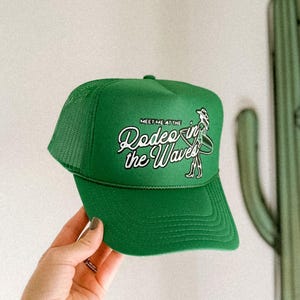 Puede incluir: Gorra de camionero verde con un logotipo blanco que dice "Meet me at the Rodeo in the Waves" con un dibujo animado de un surfista montando una ola.