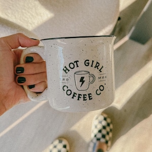 May include: A white ceramic mug with black speckles and a black logo that says "Hot Girl Coffee Co." with a lightning bolt and a small coffee cup. The mug has a black rim and handle.