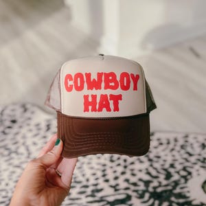 Puede incluir: Gorra de camionero marr&oacute;n y beige con las palabras "COWBOY HAT" en letras rojas. La gorra tiene una parte trasera de malla y una visera curva. La gorra est&aacute; siendo sostenida por una persona.