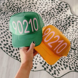 Può includere: Due cappellini da camionista, uno verde e uno arancione, ciascuno con la scritta "90210" in rosa. I cappellini sono tenuti in mano, su un tappeto maculato bianco e nero.