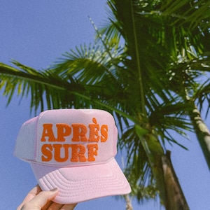 May include: Pink trucker hat with an orange "Après Surf" logo. The hat is being held up against a blue sky with palm trees in the background.