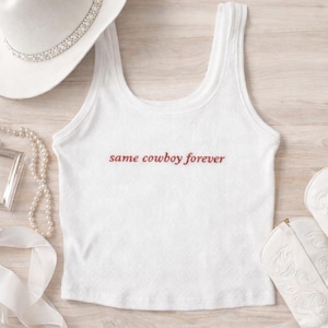 May include: White ribbed tank top with the text "same cowboy forever" in a reddish-brown font. The image also includes a white cowboy hat with a rhinestone band, white cowboy boots, and jewelry.