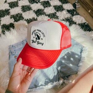 Puede incluir: Gorra de camionero roja y blanca con un parche bordado en negro que dice "Support Your Local Girl Gang" con un dibujo de una mujer con un sombrero de vaquero.