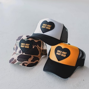 May include: Three trucker hats with the text "GIRLS JUST WANT BEER" on a black heart-shaped patch. One hat is white with a black brim, another is camouflage, and the third is orange with a black brim.