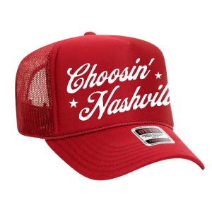 May include: Red trucker hat with white text that reads "Choosin' Nashville" with star accents. The hat has a mesh back and a curved brim. The brand label is visible on the brim.