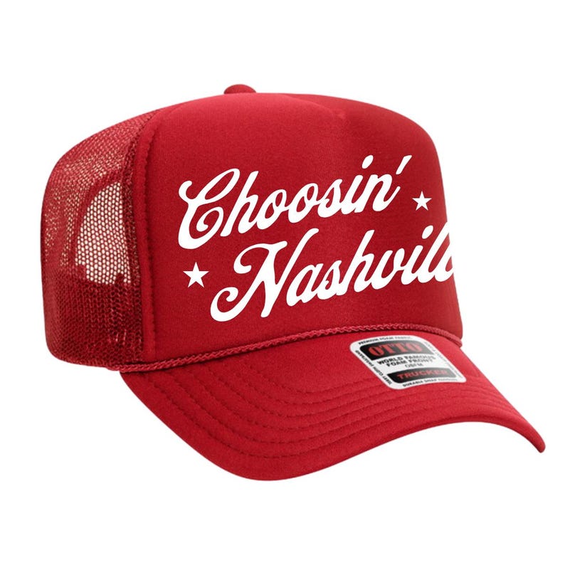May include: Red trucker hat with white text that reads "Choosin' Nashville" with star accents. The hat has a mesh back and a curved brim. The brand label is visible on the brim.