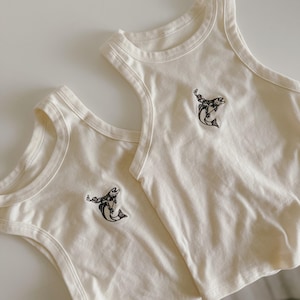 May include: Two cream-coloured tank tops featuring a black graphic of a person riding a fish. The tank tops are made of a soft material and have a classic, sleeveless design. The graphic is centered on the chest of each top.