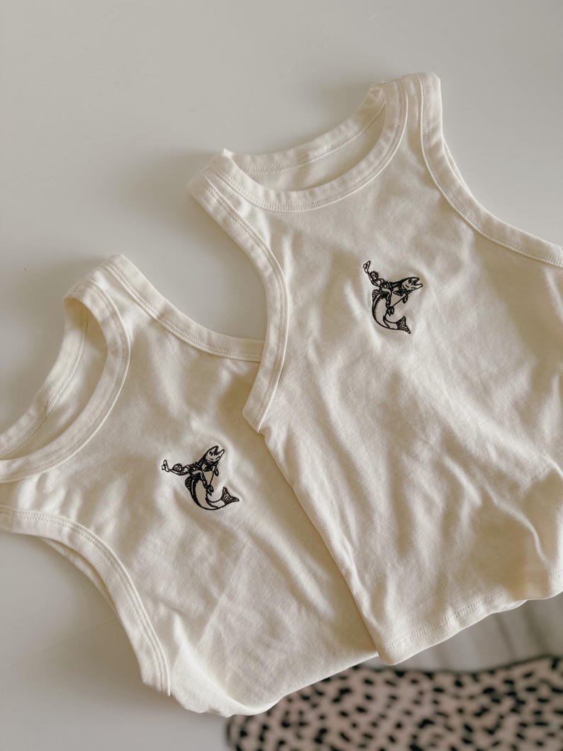 May include: Two cream-coloured tank tops featuring a black graphic of a person riding a fish. The tank tops are made of a soft material and have a classic, sleeveless design. The graphic is centered on the chest of each top.