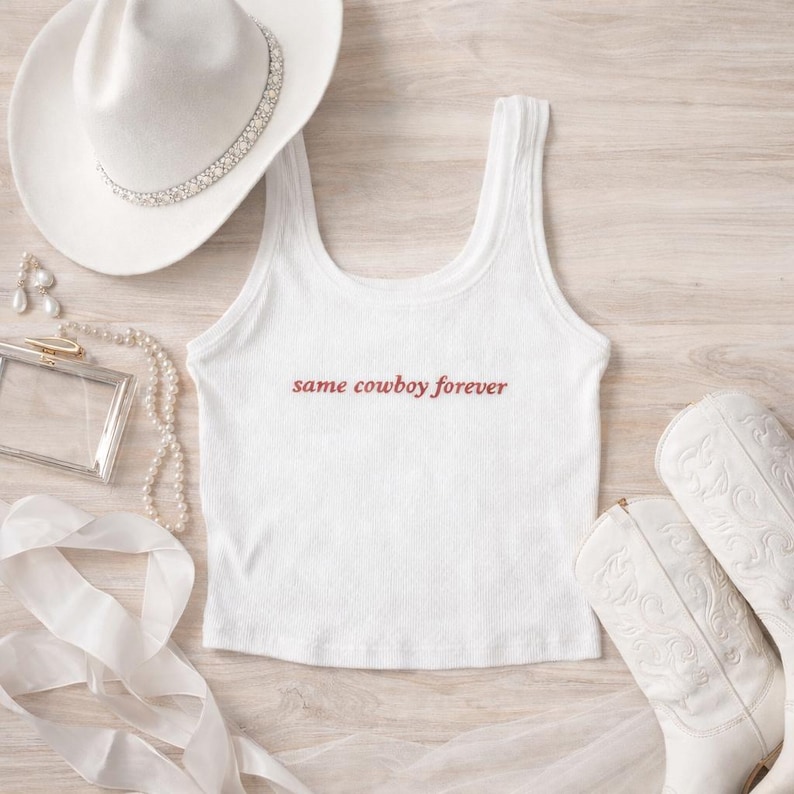 May include: White ribbed tank top with the text "same cowboy forever" in a reddish-brown font. The image also includes a white cowboy hat with a rhinestone band, white cowboy boots, and jewelry.