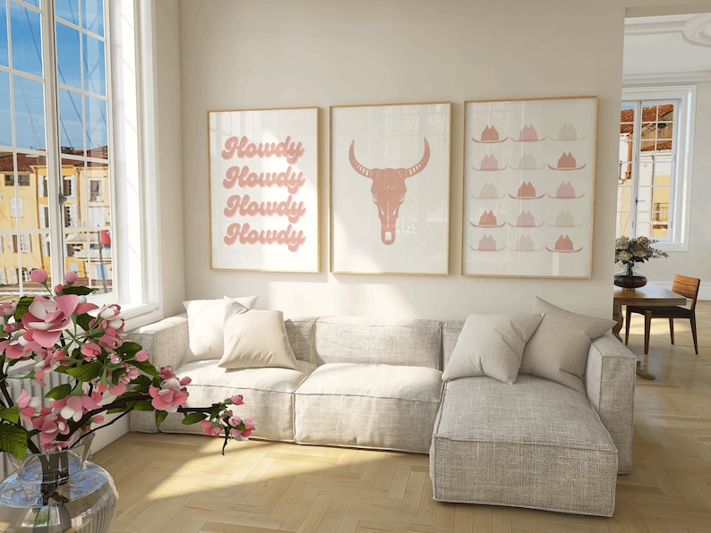 May include: Three framed prints in a living room setting. The prints are in a light pink color and feature a cowboy skull, the word "Howdy" repeated four times, and a pattern of cowboy hats.