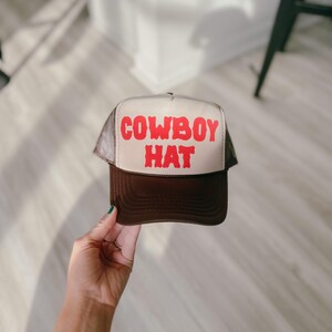 Puede incluir: Gorra de camionero marr&oacute;n y beige con la inscripci&oacute;n "COWBOY HAT" en rojo. La gorra tiene una parte trasera de malla y una visera curva. Un sombrero cl&aacute;sico de estilo occidental.