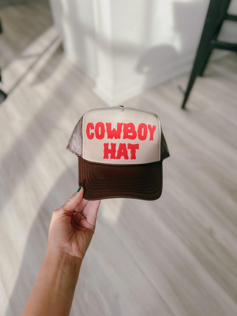 Puede incluir: Gorra de camionero marr&oacute;n y beige con la inscripci&oacute;n "COWBOY HAT" en rojo. La gorra tiene una parte trasera de malla y una visera curva. Un sombrero cl&aacute;sico de estilo occidental.